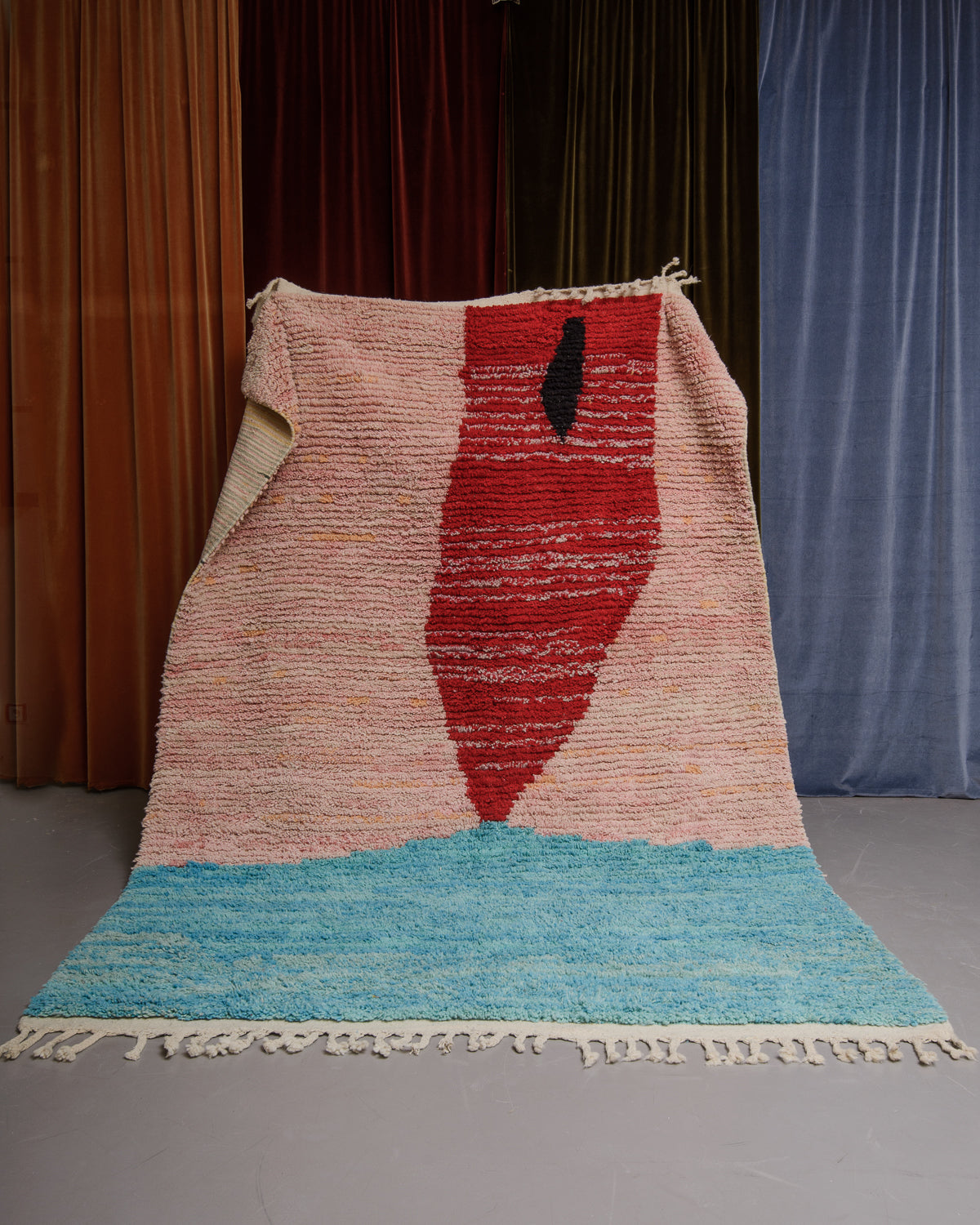 Berberlin Boujaad rug in pink, red and turquoise wool being held in front of velvet curtains.