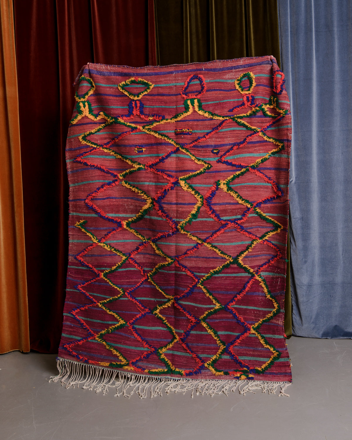 Vintage flat woven Kelim with fluffy embroidered pattern, held in front of velvet curtains. 