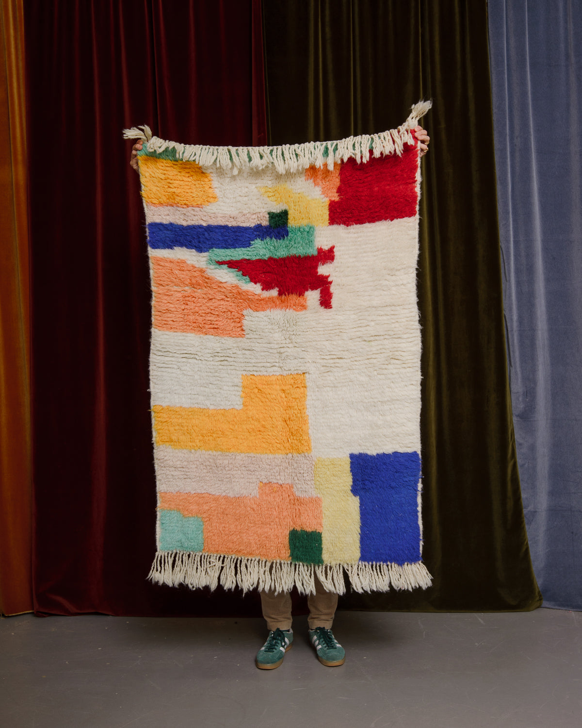 Colourful wool rug being held up in front of velvet curtains.