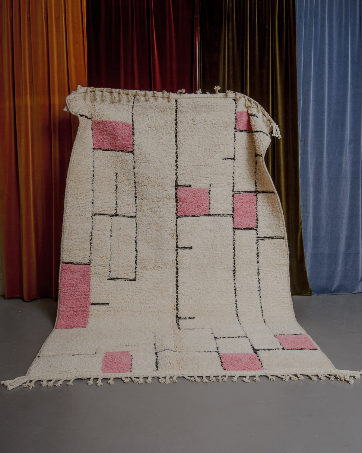 Cream, pink and black Berberlin Beniourain rug held up in front of velvet curtains.