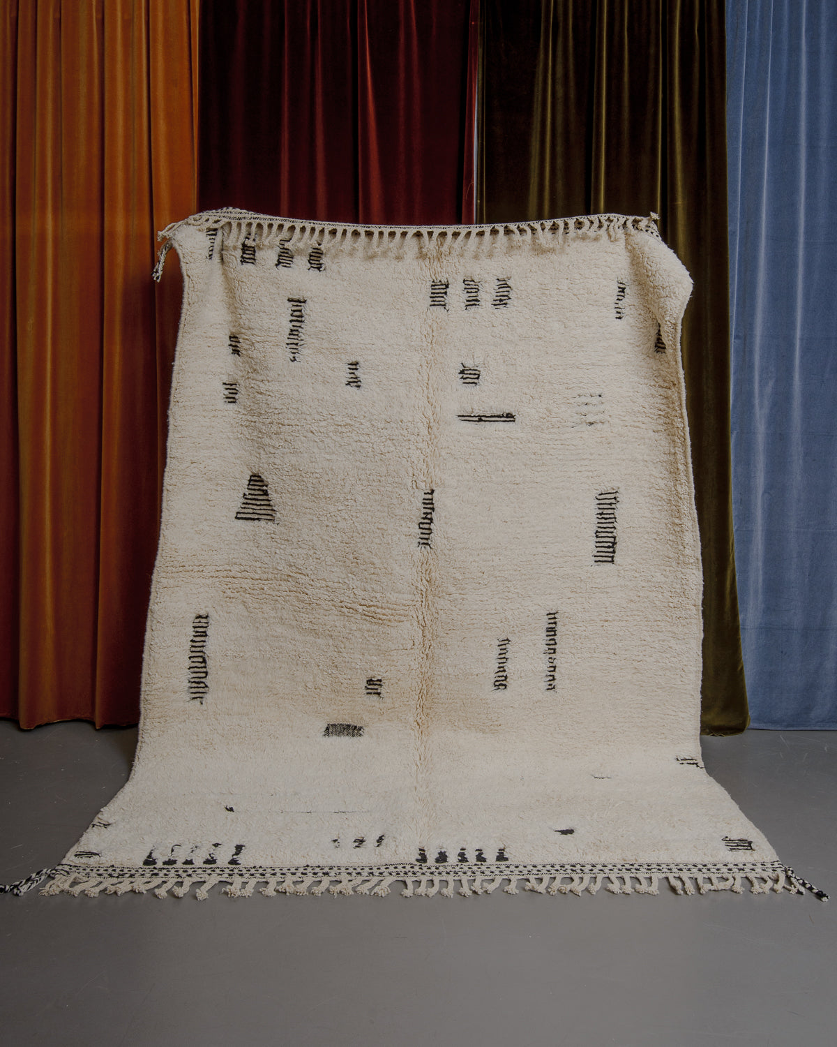 Cream and black Berberlin Beniourain rug held up in front of velvet curtains.