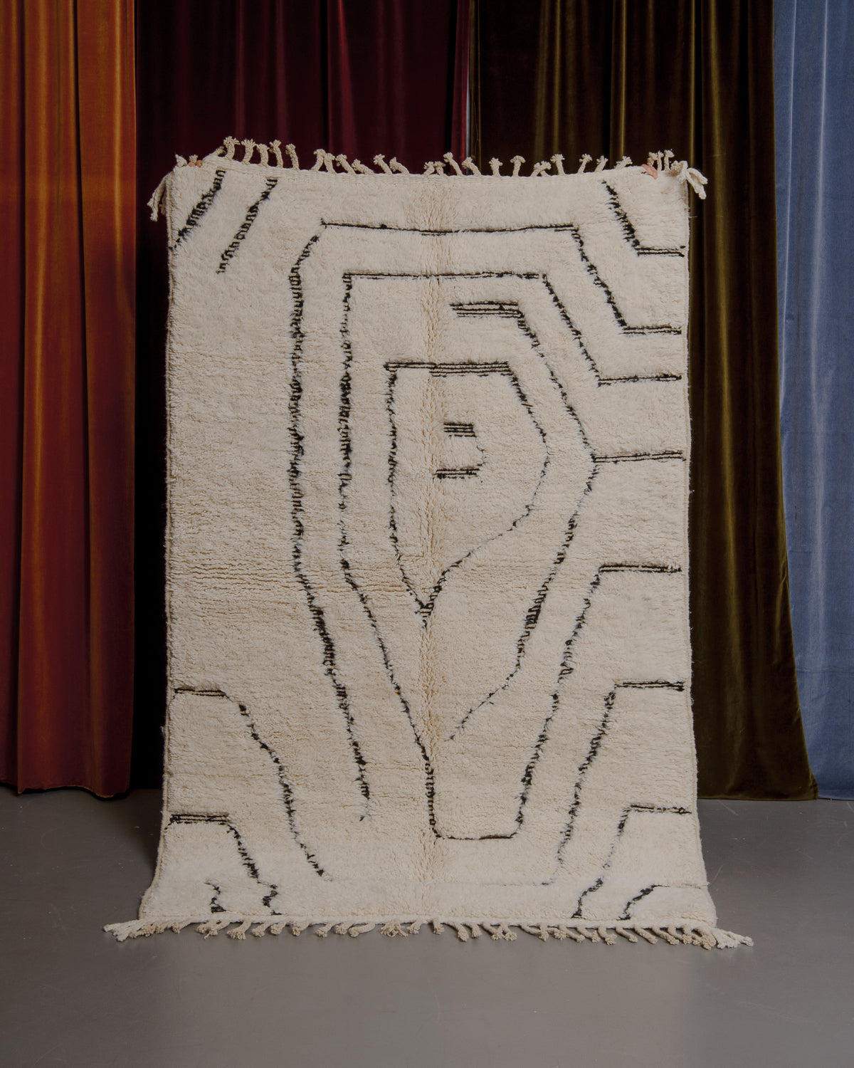 Cream and black Berberlin Beniourain rug held up in front of velvet curtains.