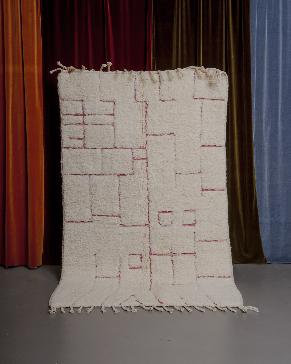 Cream and pink Berberlin Beniourain rug held up in front of velvet curtains.