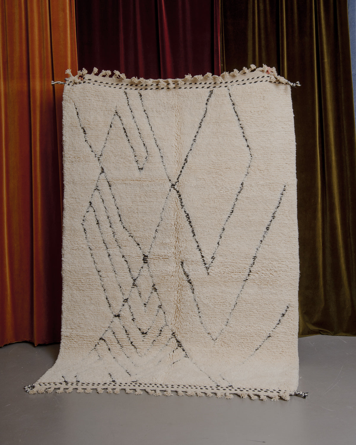 Cream and black Berberlin Beniourain rug held up in front of velvet curtains.