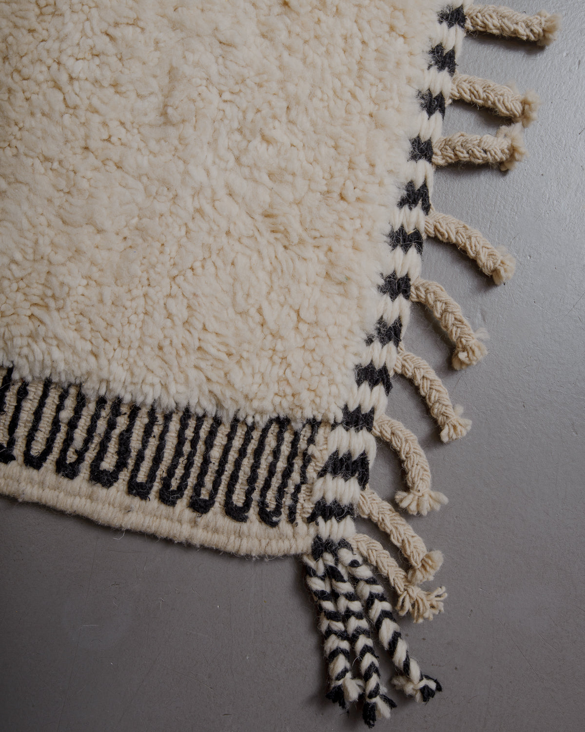 Corner of cream beniourain rug with natural wool tassels and black and white pattern along the end and sides.