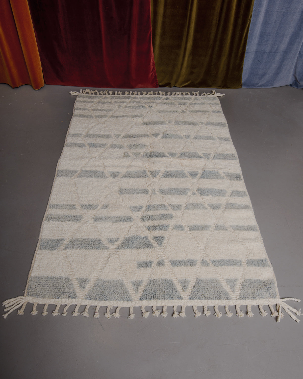 Cream and light blue-grey Berberlin Beniourain rug on grey floor in front of velvet curtains.