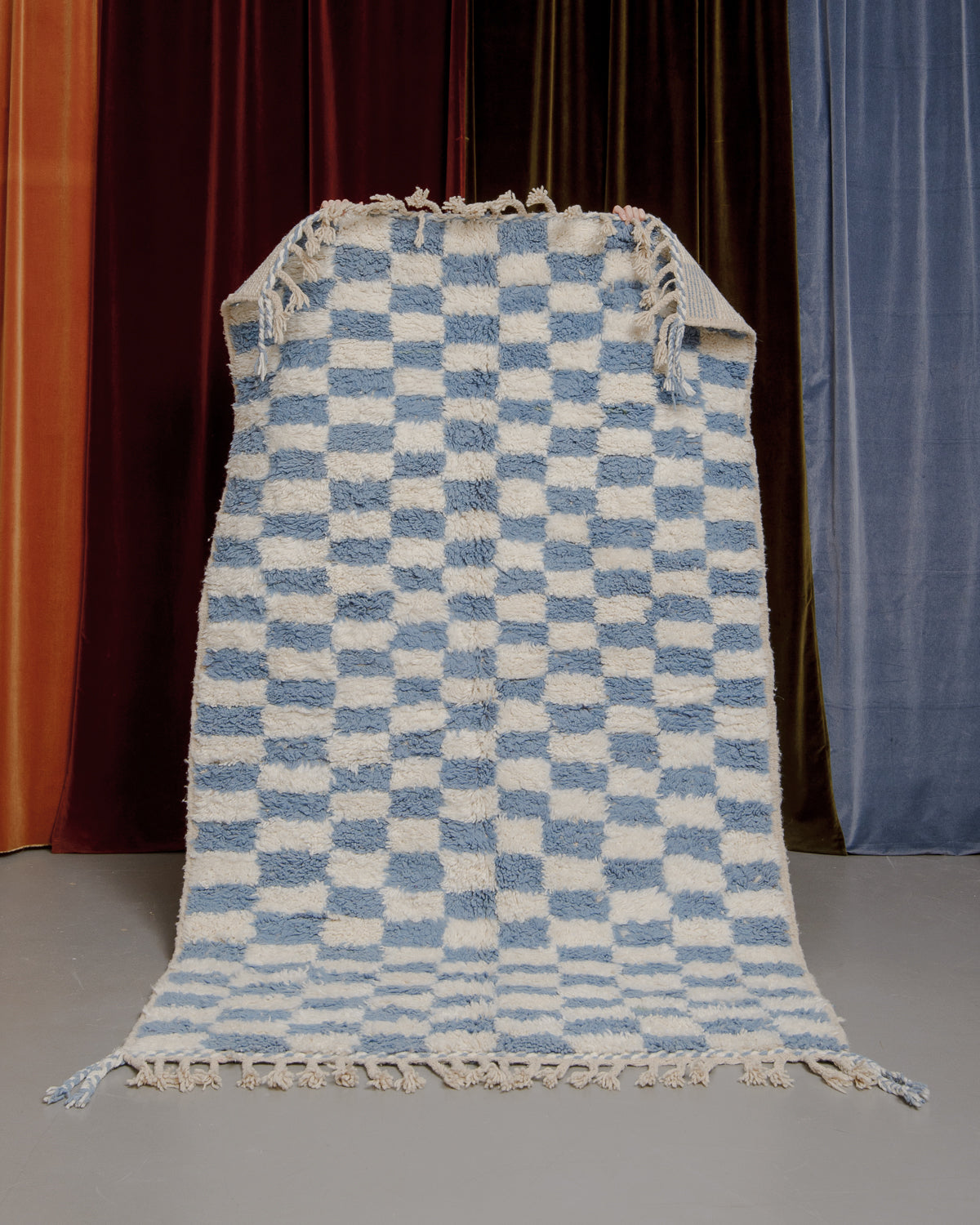 Berberlin Beniourain rug with blue and cream checkered pattern being held up in front of velvet curtains.