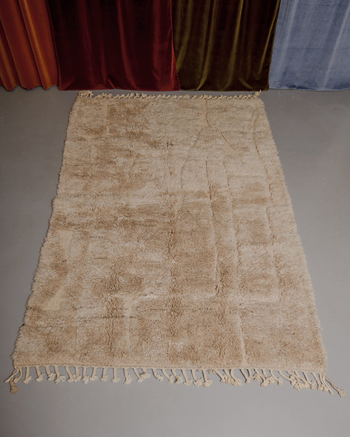 Beige brown fluffy Berberlin Beniourain rug on grey floor in front of velvet curtains.