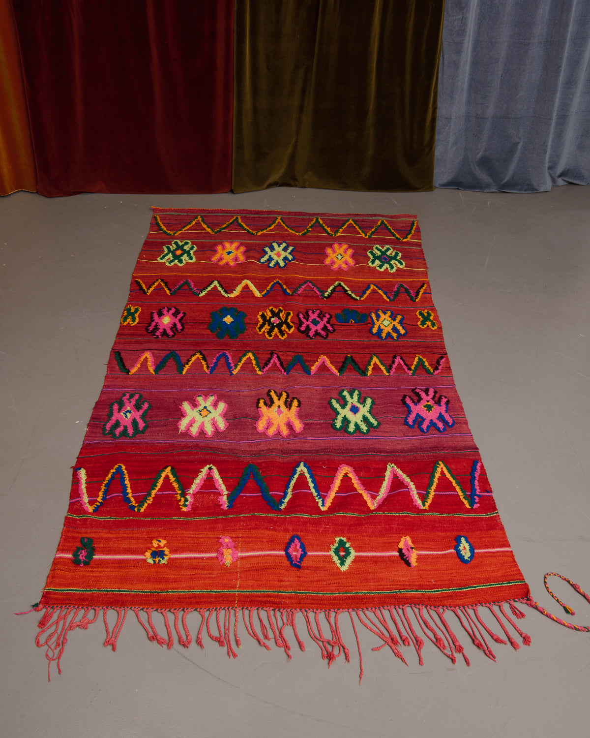 Vintage flat woven Kelim with fluffy embroidered pattern, on grey floor in front of velvet curtains. 