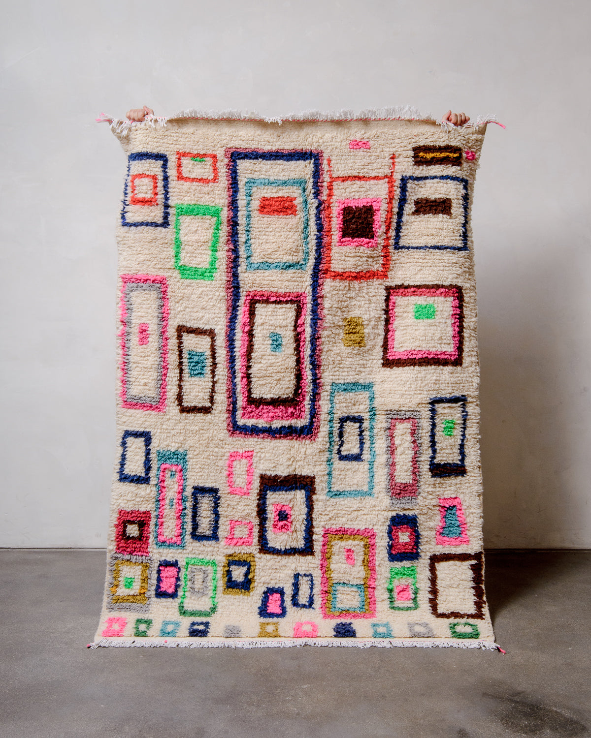 Berberlin Azilal rug with colourful design held up by hands in front of white studio wall. 