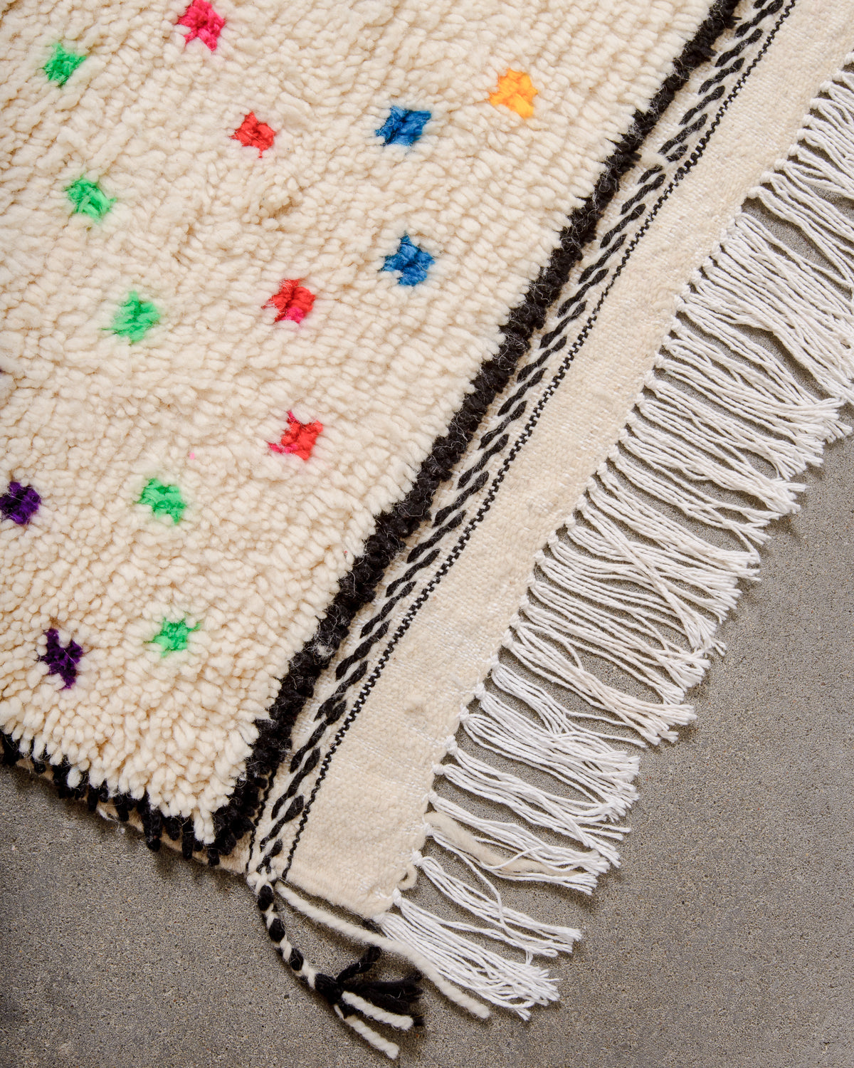 Corner of Azilal rug with white cotton tassels and a black border, laying on concrete floor. 