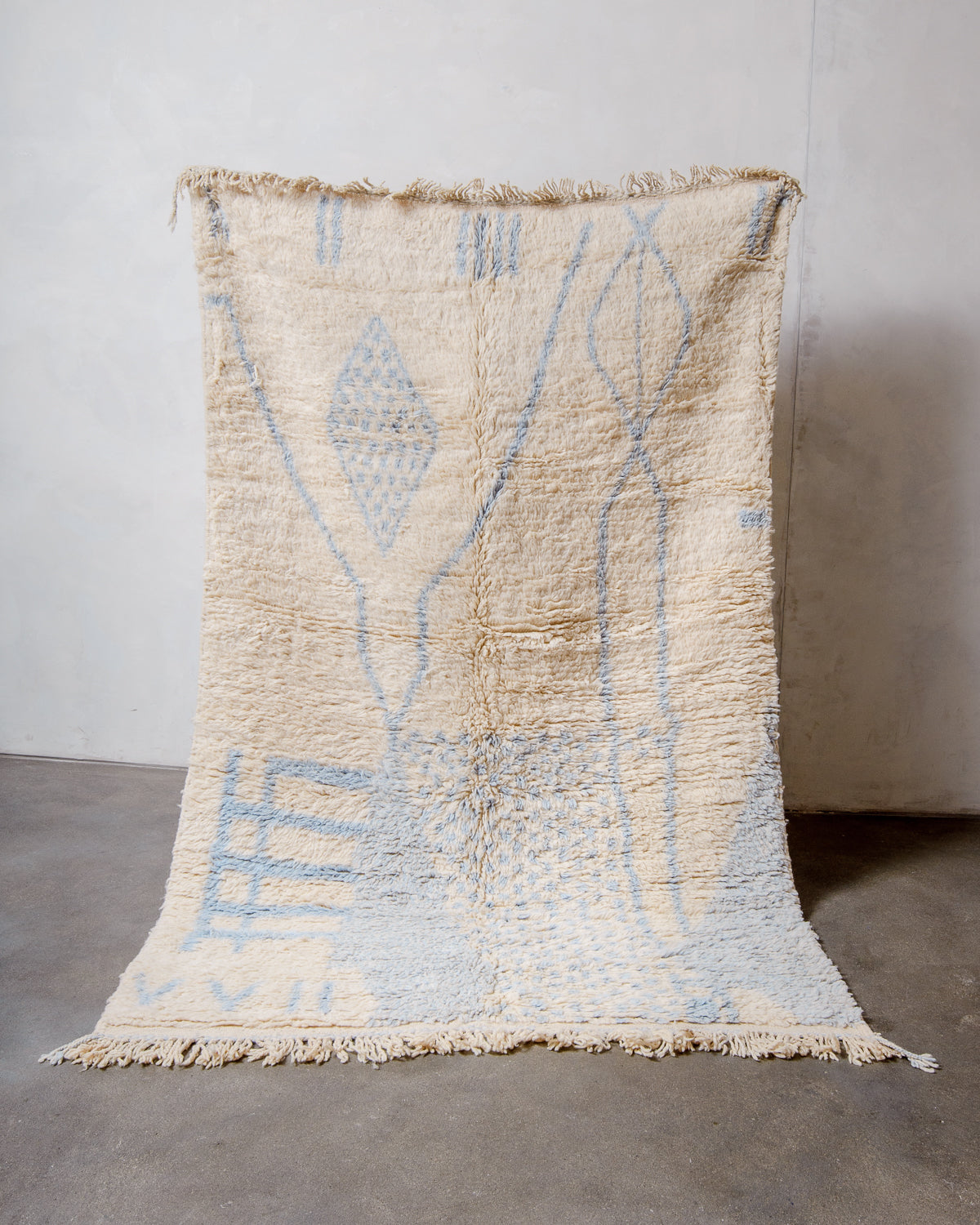 Berberlin Azilal rug in fluffy cream and light blue wool, being held up in front of a white wall and grey concrete floor. 