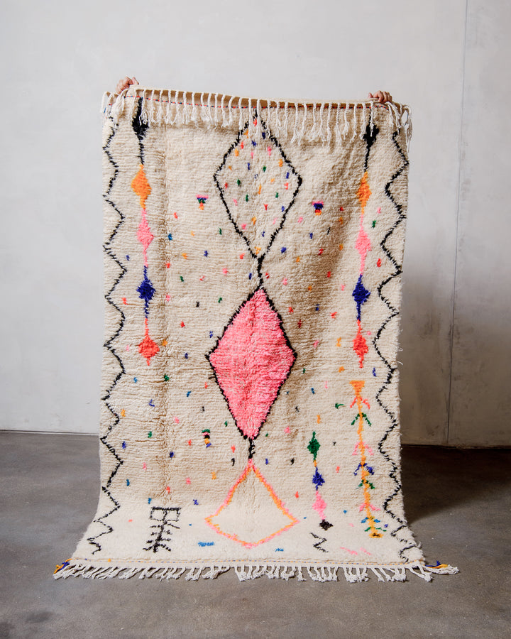 Berberlin Azilal rug made of fluffy cream wool with cotton tassels, held up  against a white wall and cement floor.