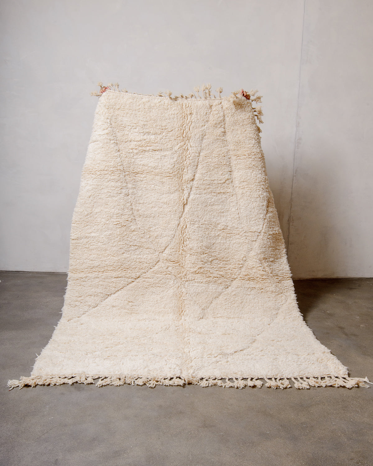 Cream wool Beniourain rug being held up in front of a white wall and concrete floor. 