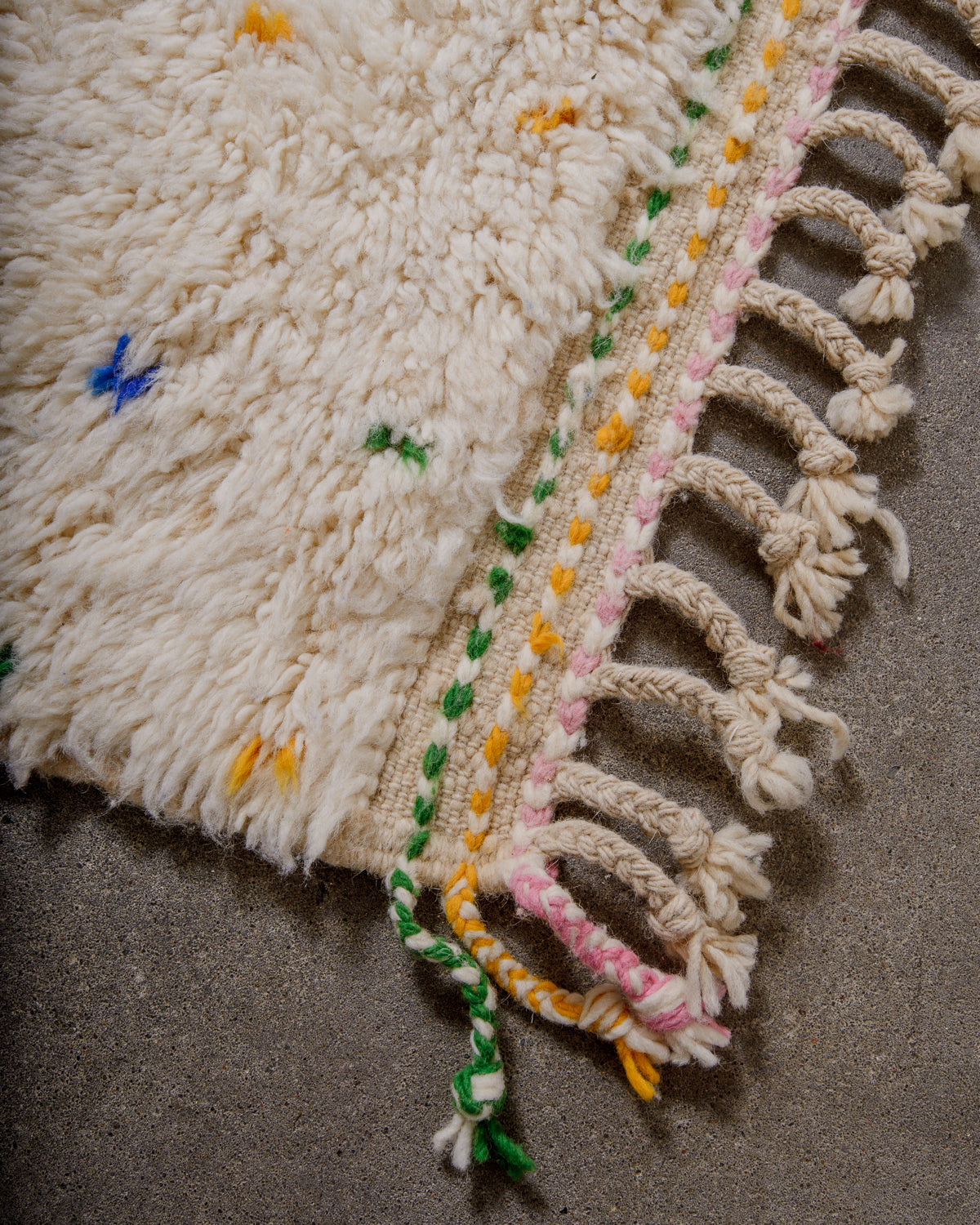 Corner of Berber rug with green, pink and orange tassels at the corner and natural wool tassels across the top. 