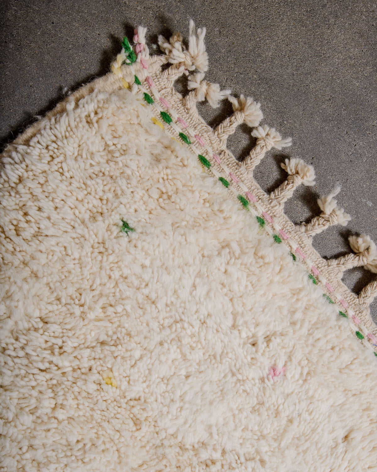 Corner of Berber rug with green, pink and yellow stitching and natural wool tassels across the top.