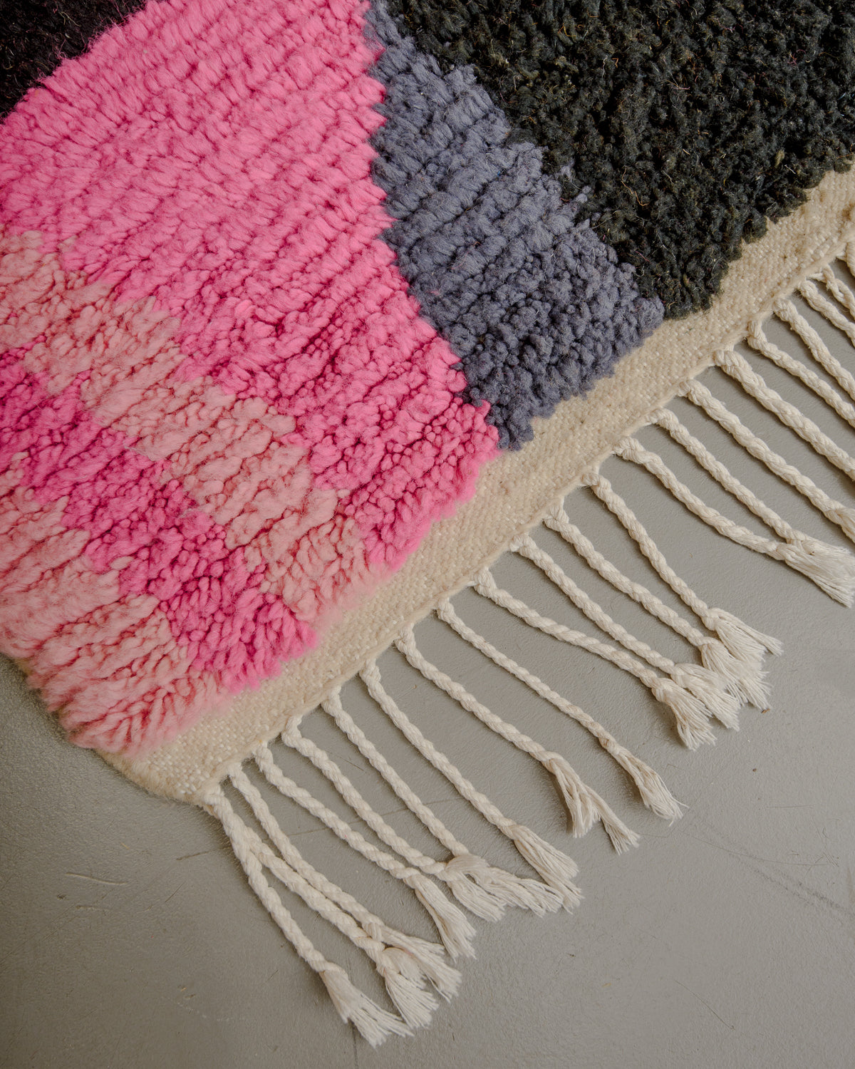 Corner of pink and grey wool rug with thin, cotton tassels. 