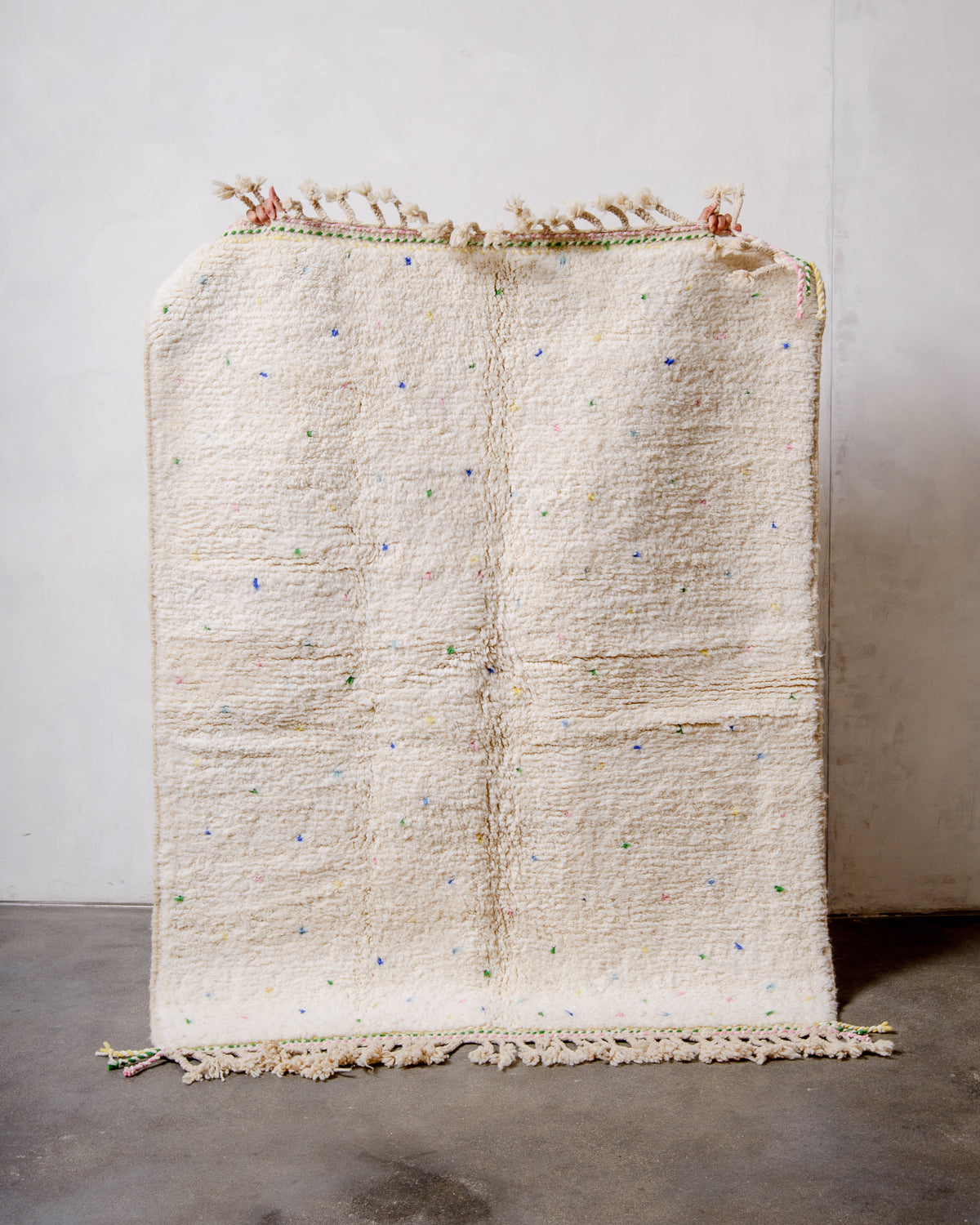 Berberlin Beniourain rug in cream wool with colourful dots being held up in front of a white wall. 