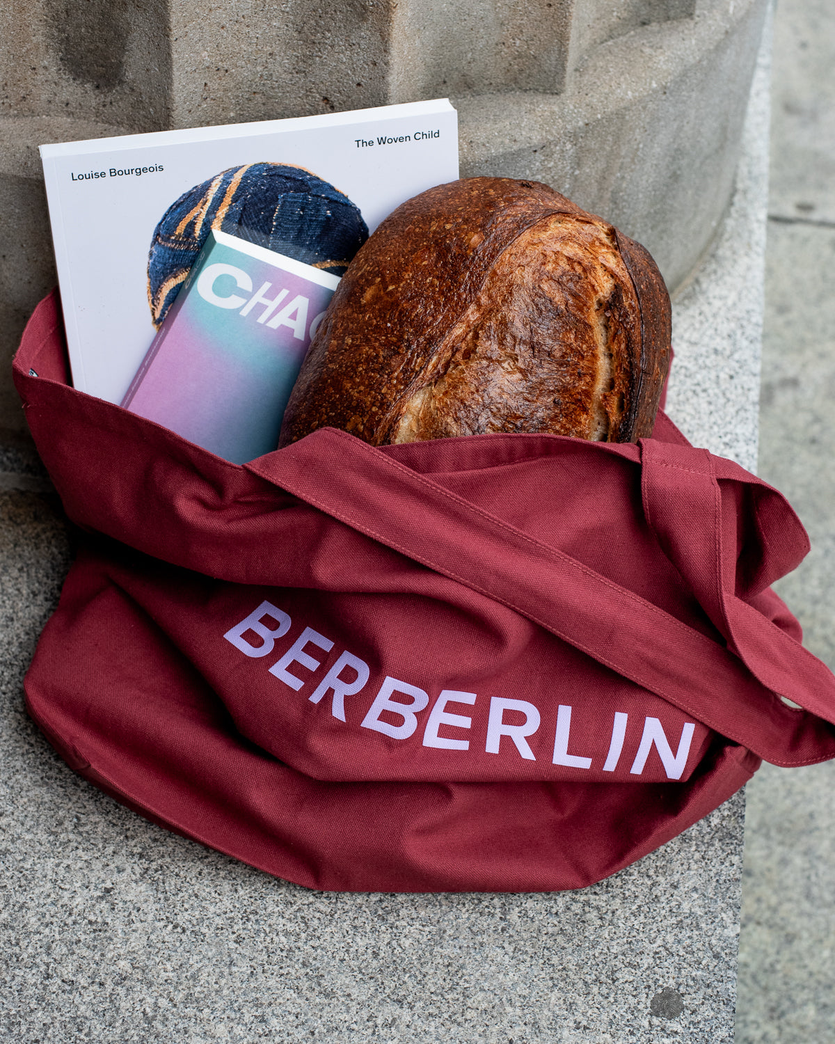 Burgundy tote bag with lilac Berberlin logo leaning against concrete column. The large bag is holding handmade bread and books. 