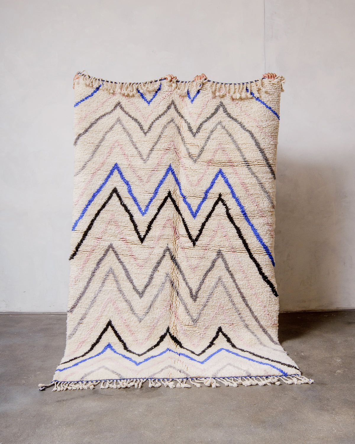 Berberlin Finest Rug made of cream wool with a pink, black, grey and blue design being held up in front of a white wall and concrete floor. 