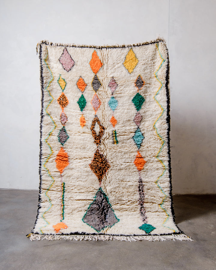 Berberlin Finest Rug made of plush cream wool with a colourful design being held up in front of a white wall and concrete floor. 