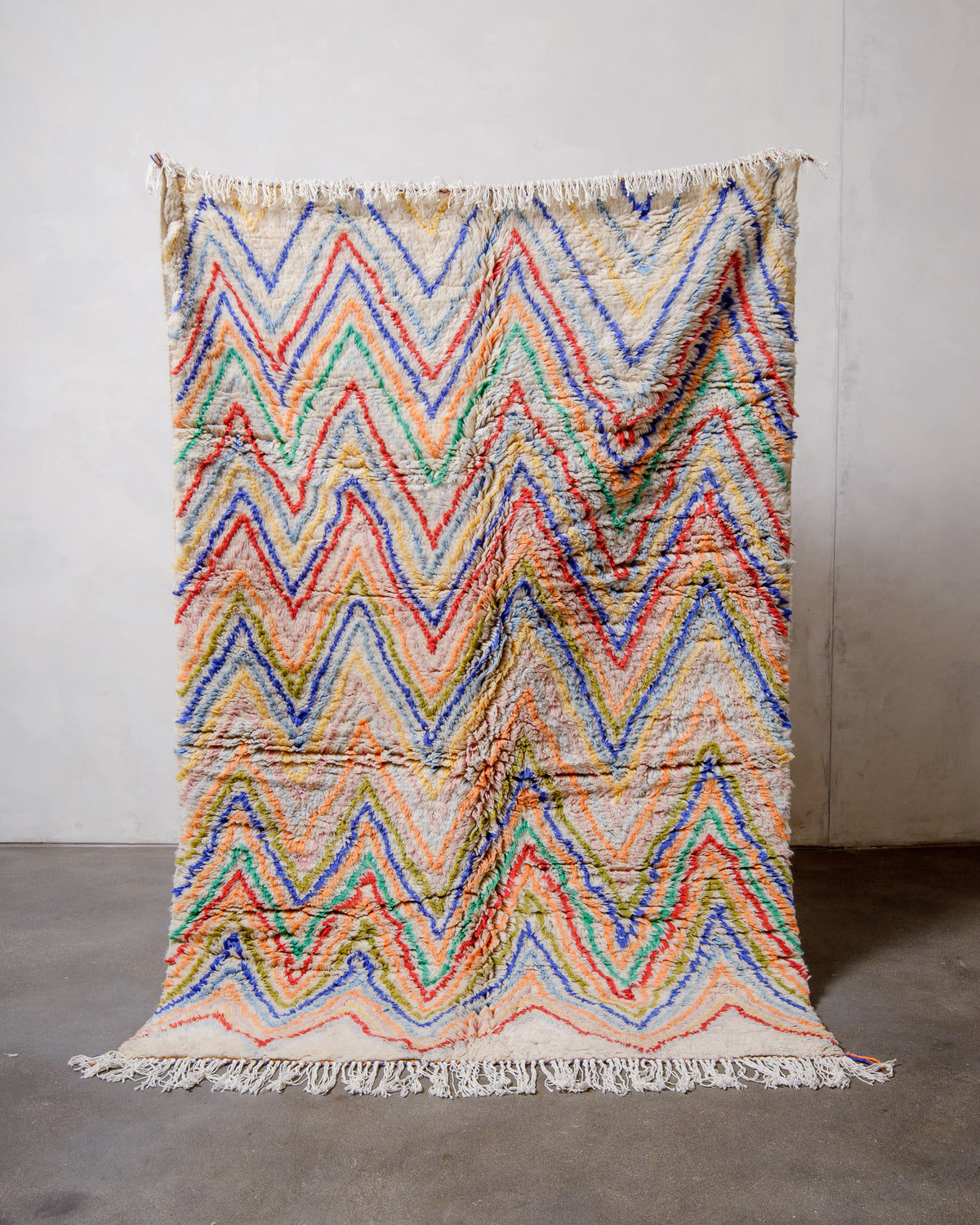 Berberlin Finest Rug made of plush cream wool with a colourful zigzag design being held up in front of a white wall and concrete floor. 