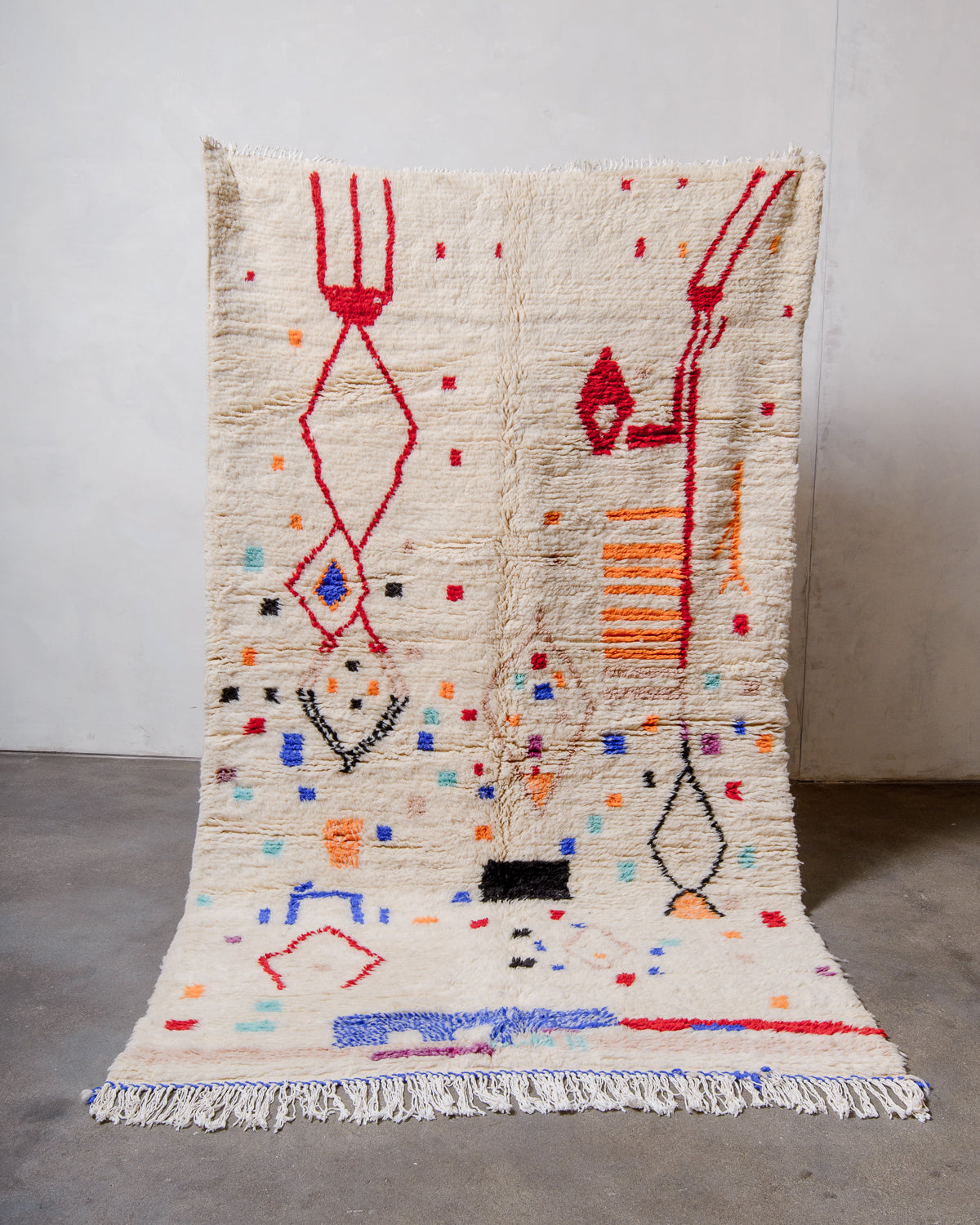 Berberlin Finest Rug made of plush cream wool with a design in red, orange, blue and black being held up in front of a white wall and concrete floor. 