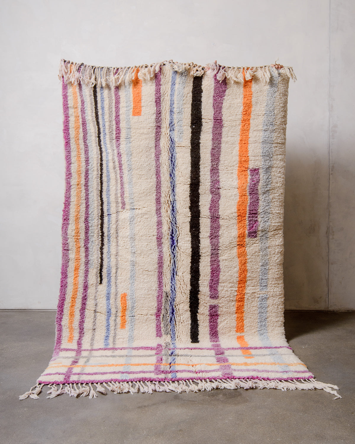 Berberlin Finest Rug made of heavy wool pile in bright colours being held up in front of a white wall and concrete floor. 