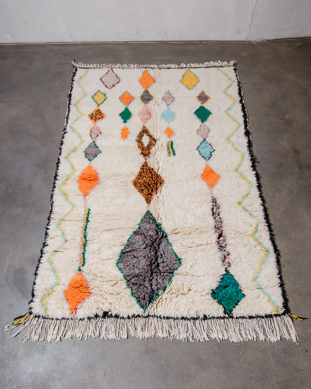 Berberlin Finest Rug made of plush cream wool with a colourful design and black border, laying flat on concrete floor. 