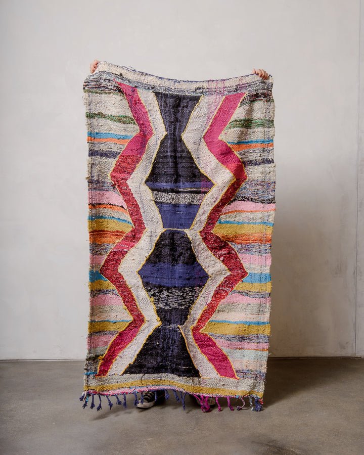 Berberlin vintage kelim rug made of recycled materials being held up by two hands in front of a white wall and concrete floor. 