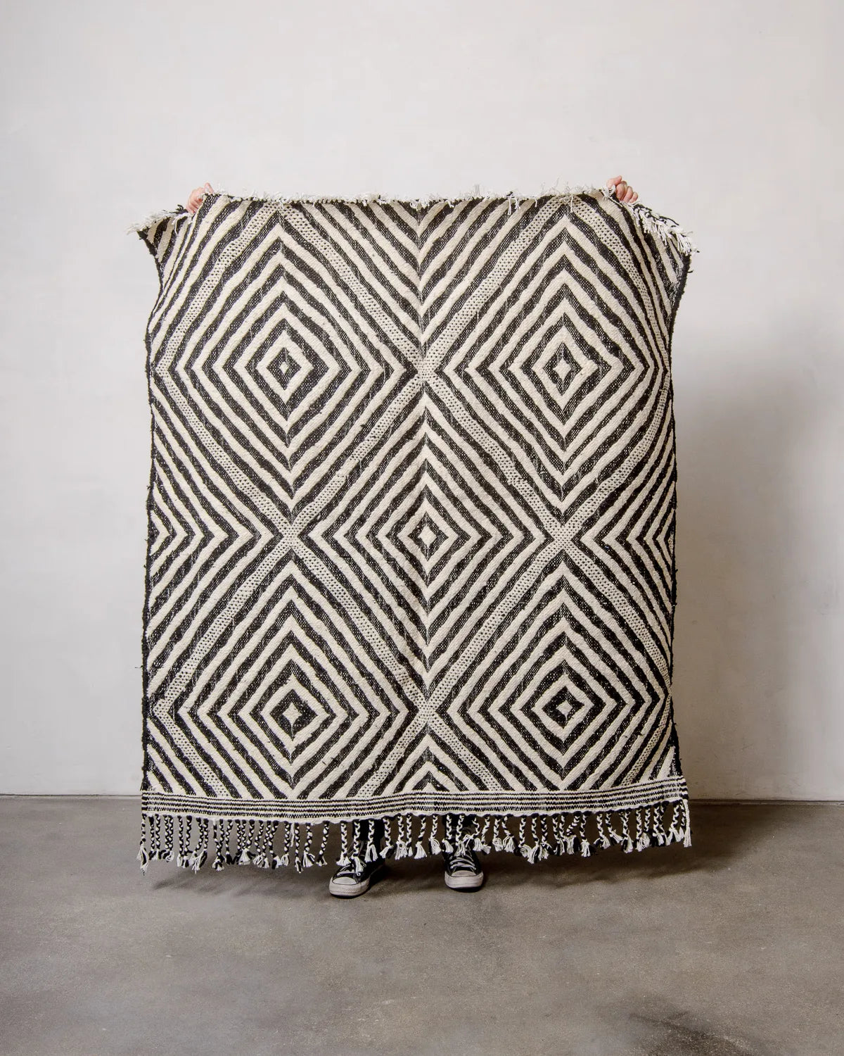Berberlin Kelim Rug in black and white, being held up in front of white wall. 