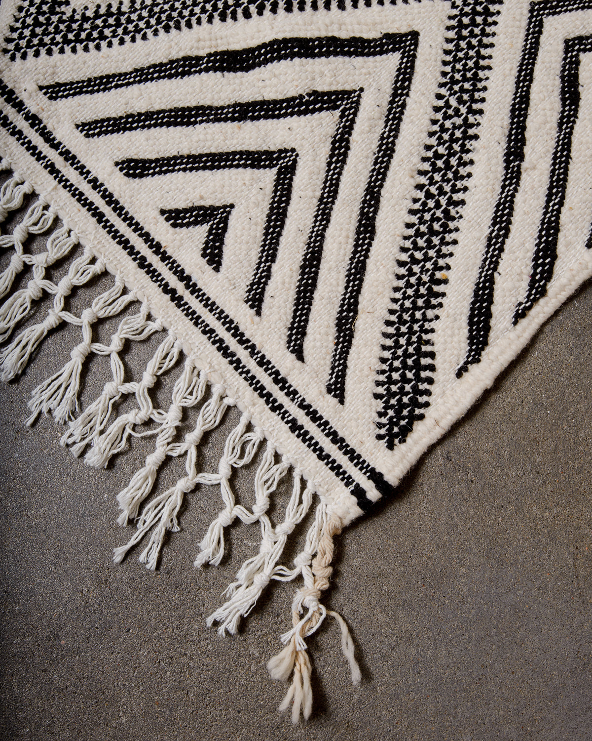 Corner of Kelim Rug in black and white with white cotton tassels, laying on concrete.