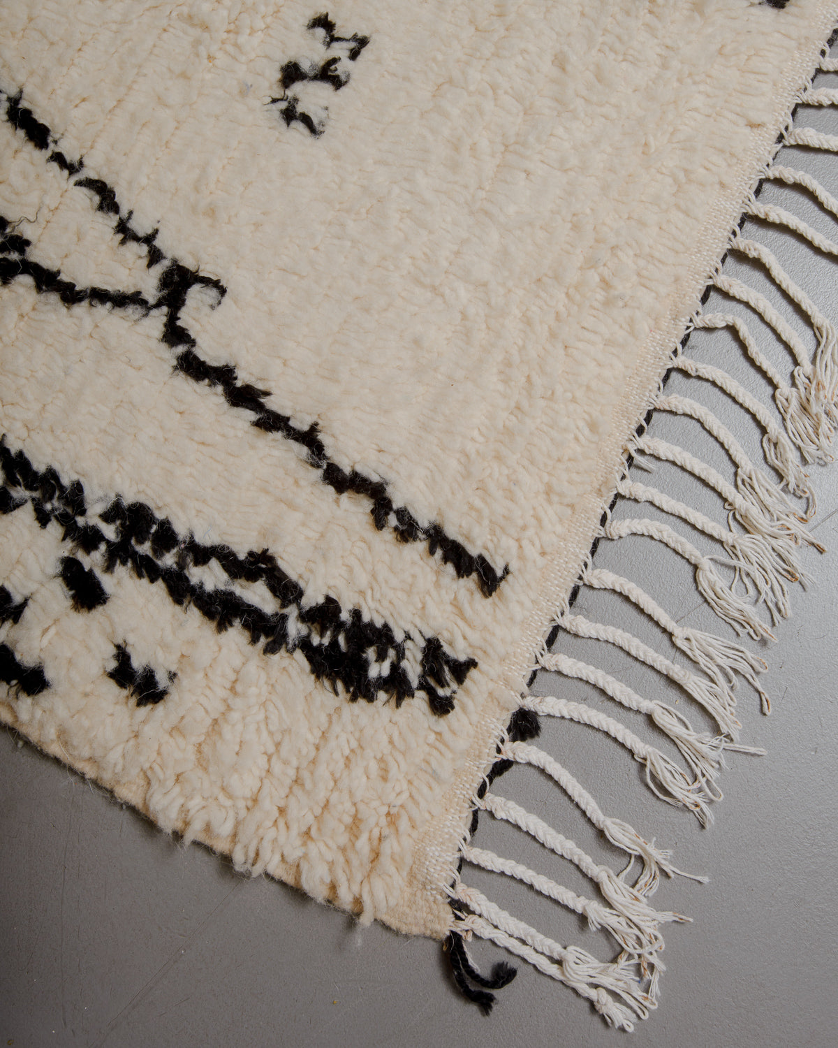 Corner of azilal rug with white cotton tassels on grey floor. 