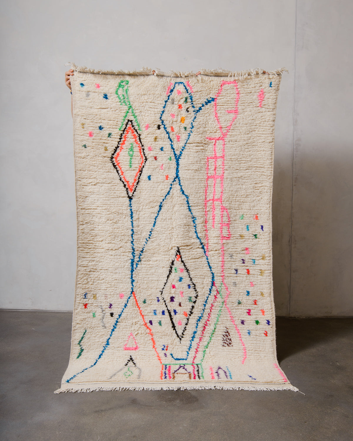 Berberlin Azilal rug made of fluffy cream wool and colourful design, held up in front of a white wall and cement floor.