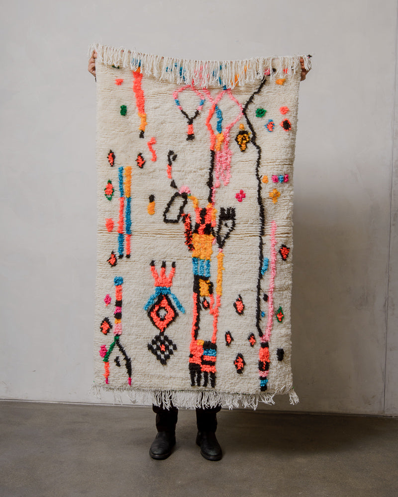 Brightly coloured Berberlin azilal rug in a small size, being held up in front of a plain wall. 