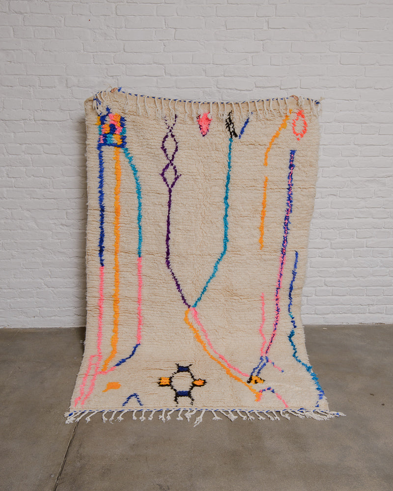 Cream Azilal rug with bold abstract design in pink, black, blue, yellow and purple being held up in front of a white brick wall. 