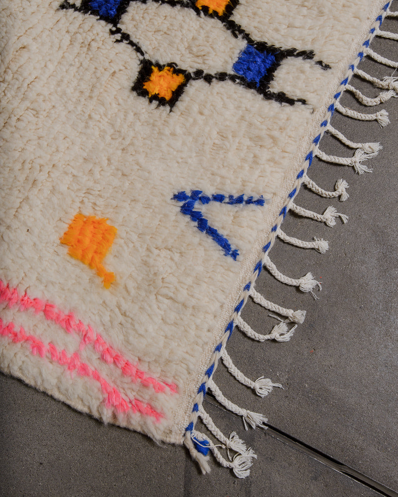 Corner of cream azilal rug with blue trim and white cotton tassels, on concrete floor. 