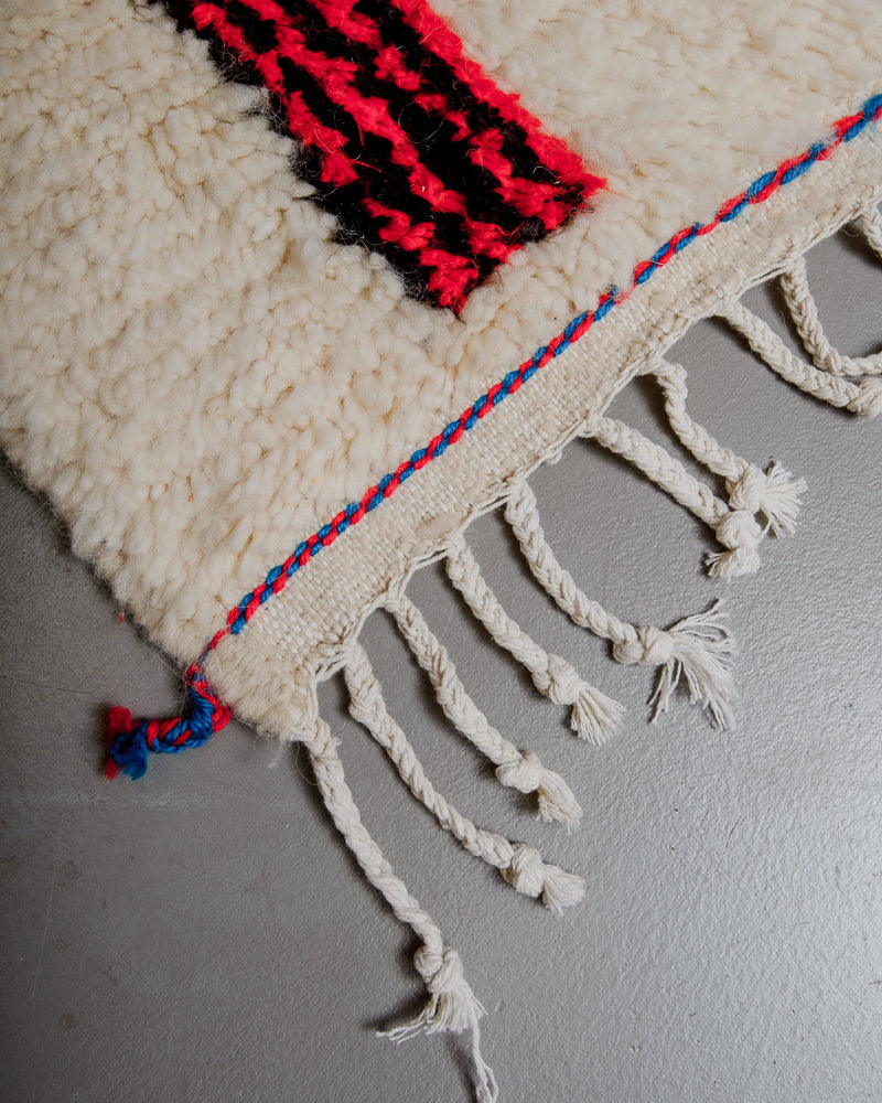 Corner of azilal rug with red and blue stitching across the end and white cotton tassels. 
