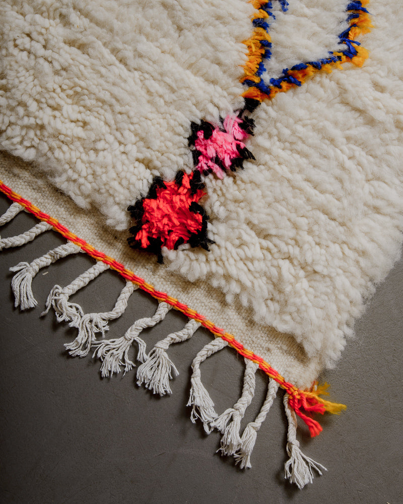 Corner of azilal rug with orange and yellow stitching along the end and white cotton tassels. 