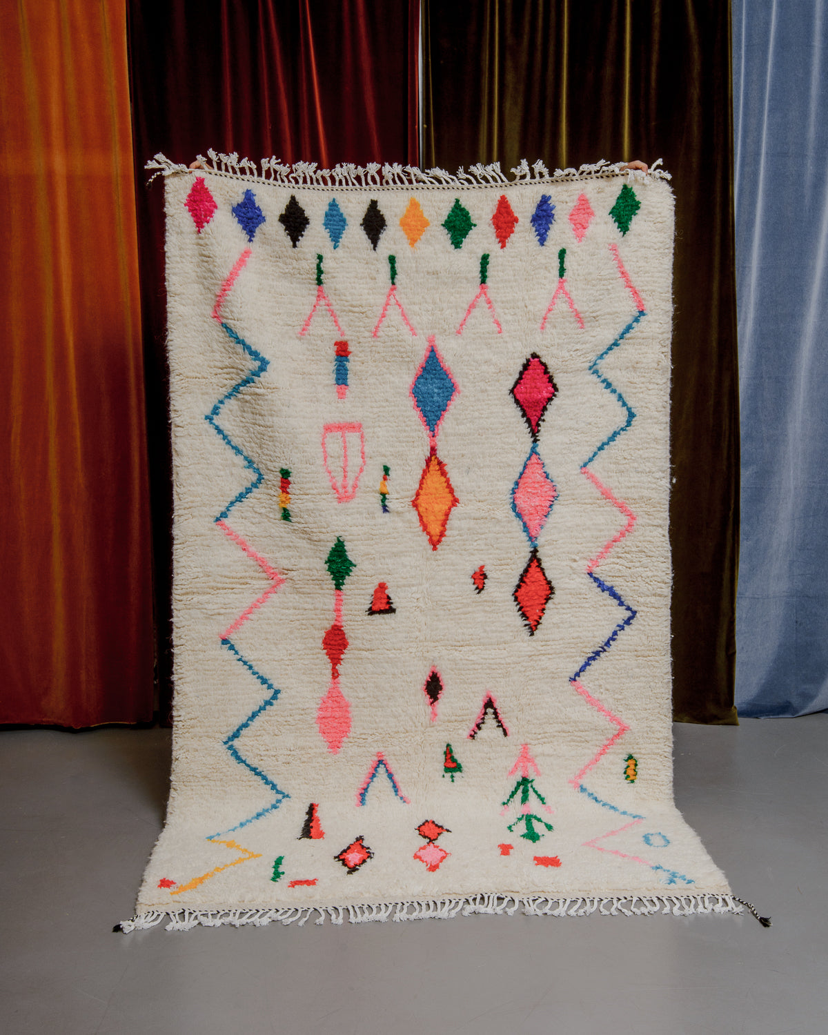 Colourful azilal rug held in front of velvet curtains. 