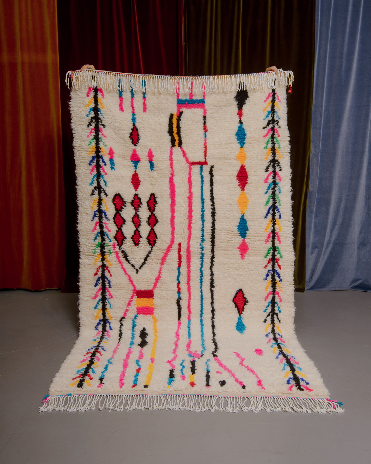 Azilal rug with colourful, abstract design being held up in front of a wall of velvet curtains.
