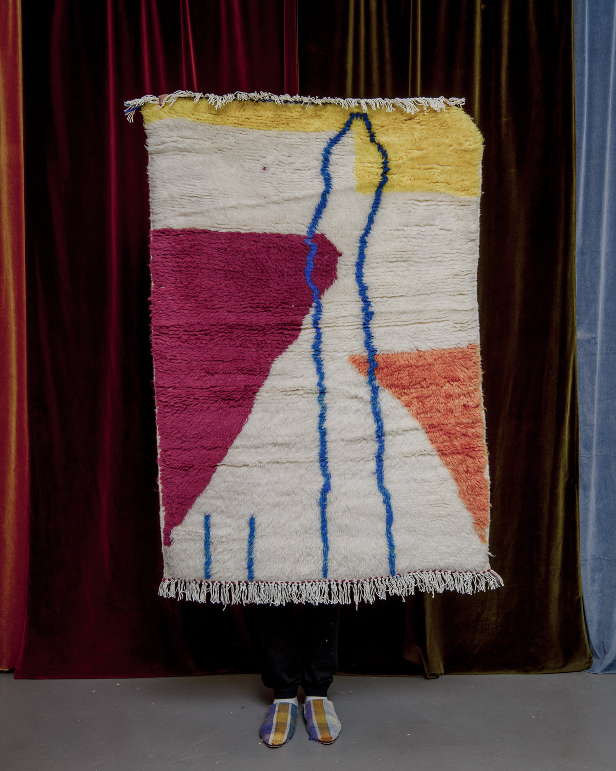 Brightly coloured Berberlin azilal rug in a small size, being held up in front of a velvet curtain.