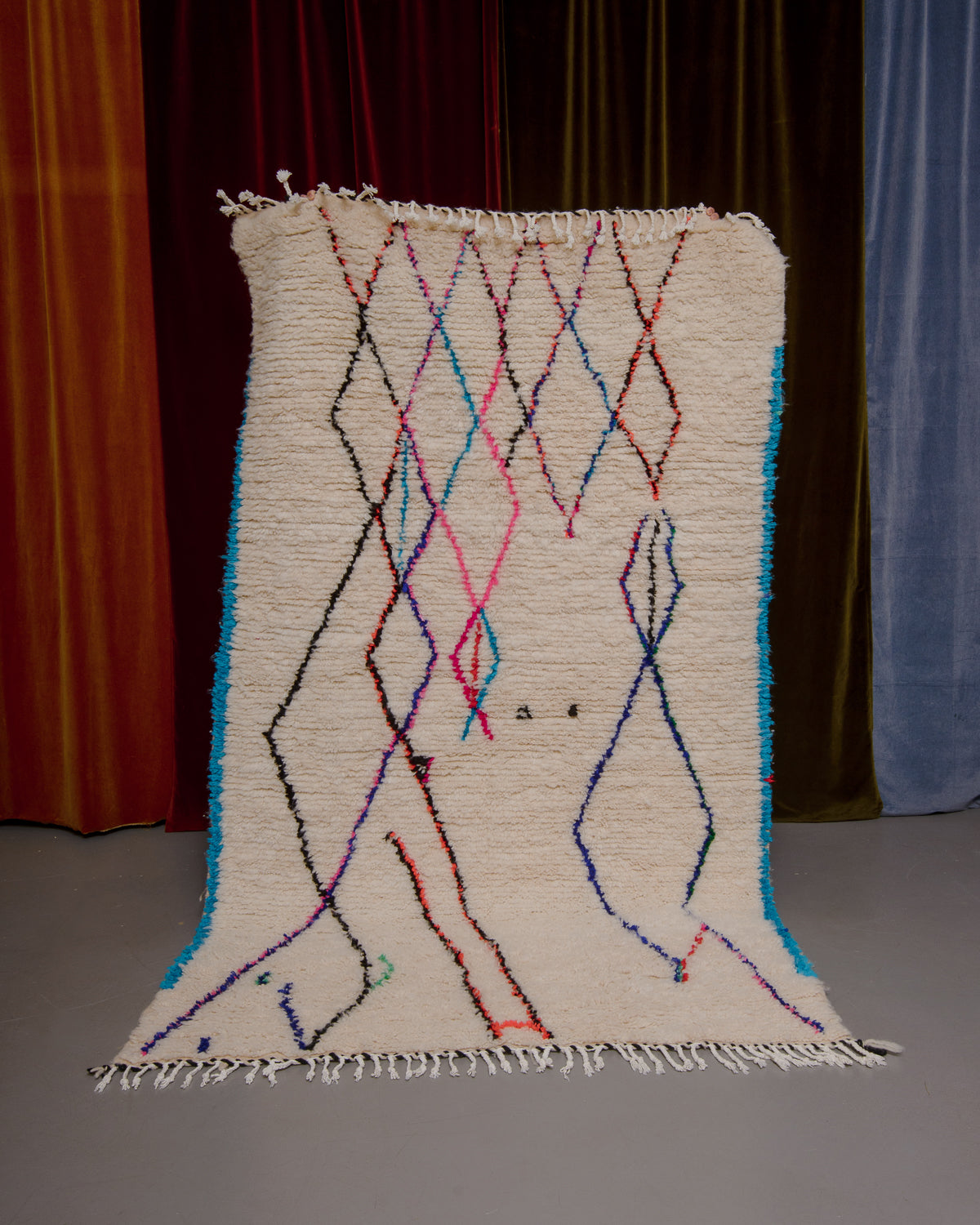 Azilal rug with colourful, abstract design being held up in front of a wall of velvet curtains.