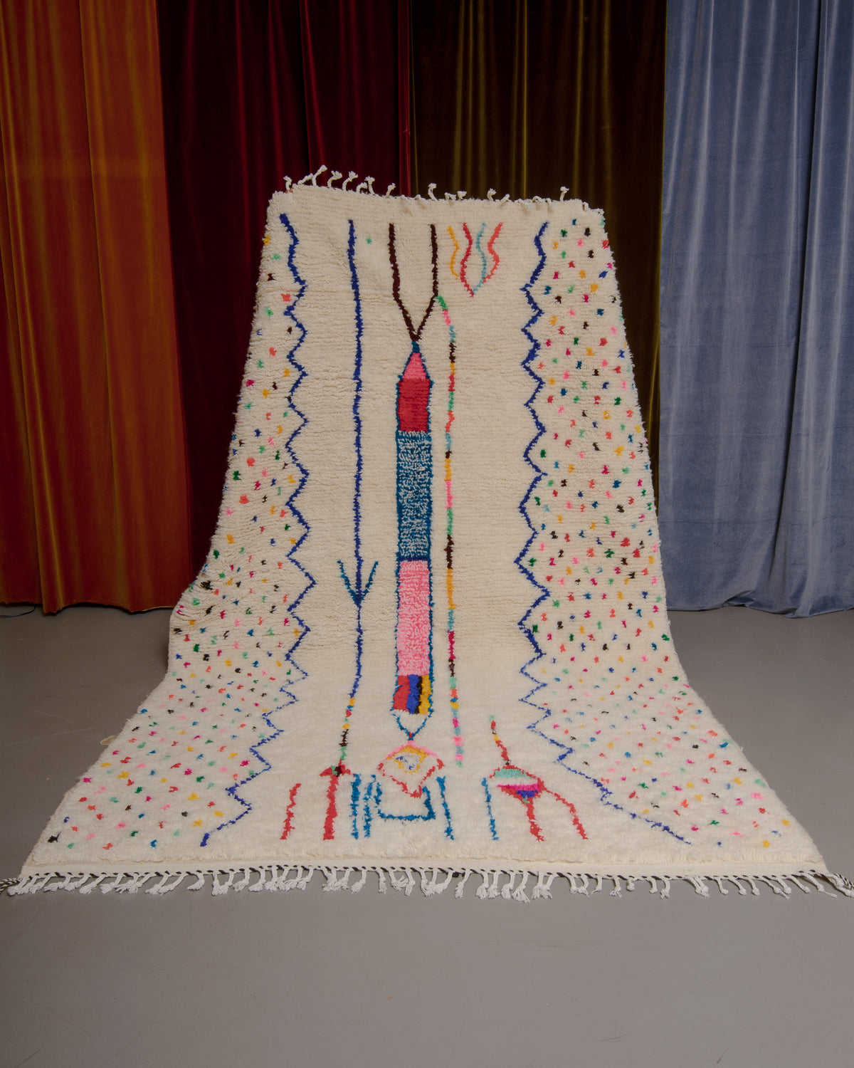 Azilal rug with colourful, abstract design being held up in front of a wall of velvet curtains.