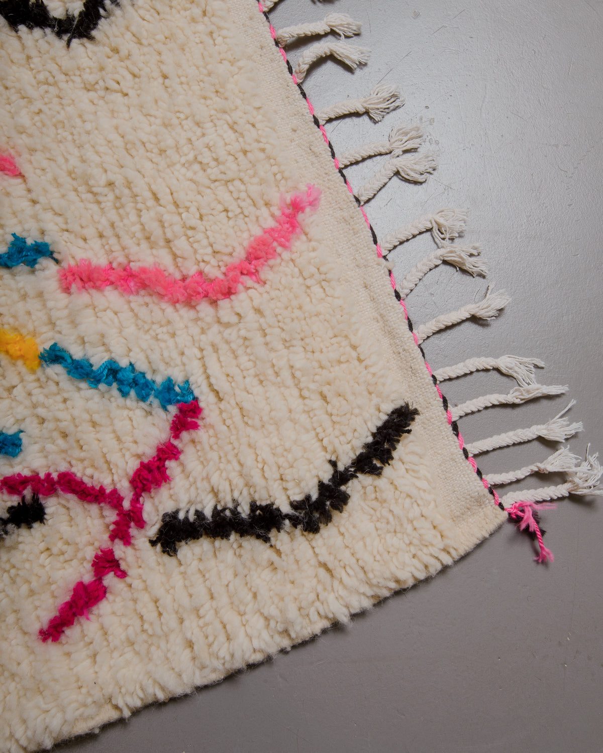 Corner of azilal rug with white cotton tassels and black and pink stitching across the end. 