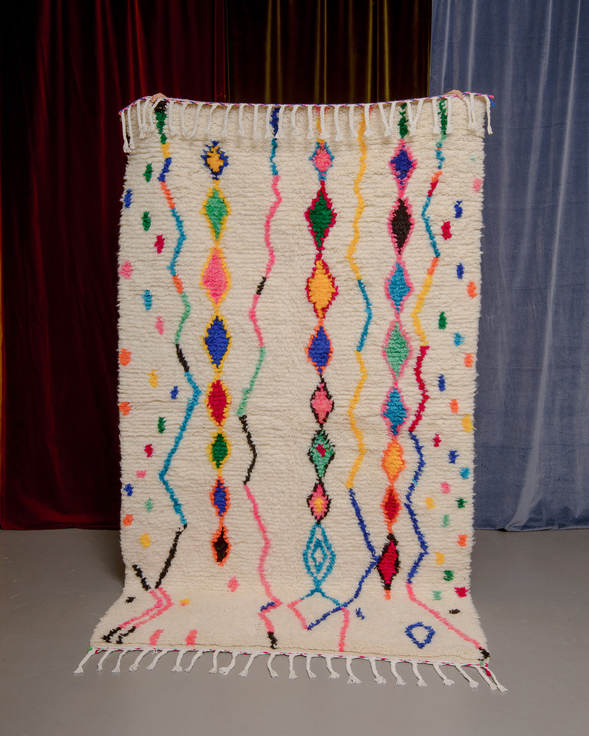 Azilal rug with colourful, abstract design being held up in front of a wall of velvet curtains.