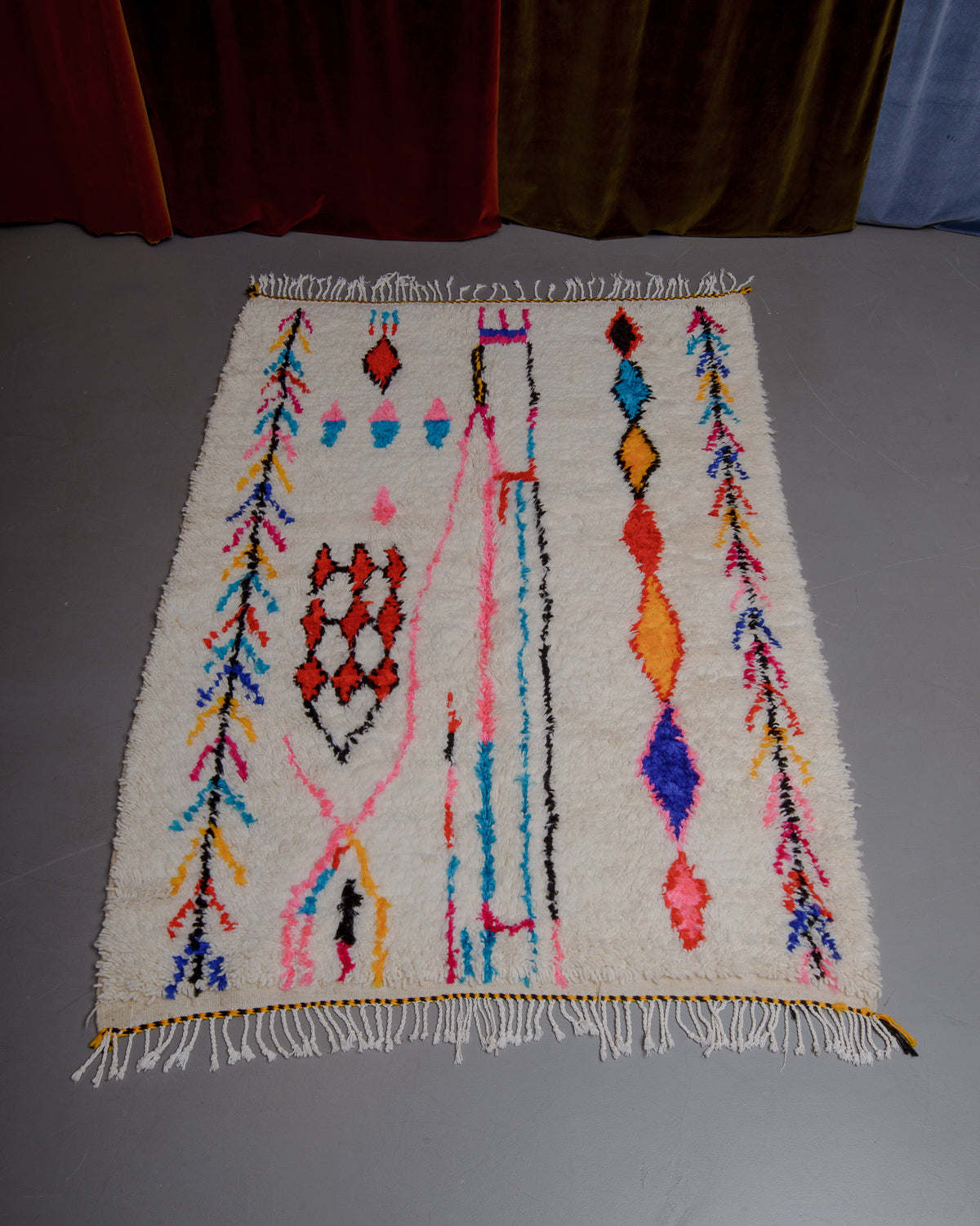 Azilal rug with colourful, abstract design laid flat on grey floor in front of a wall of velvet curtains.