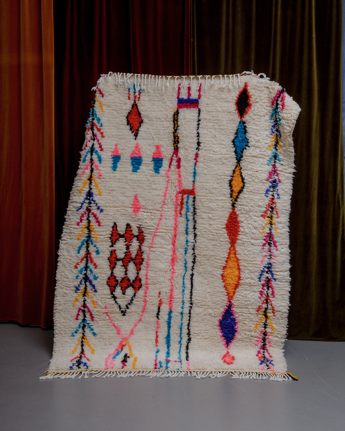 Azilal rug with colourful, abstract design being held up in front of a wall of velvet curtains.