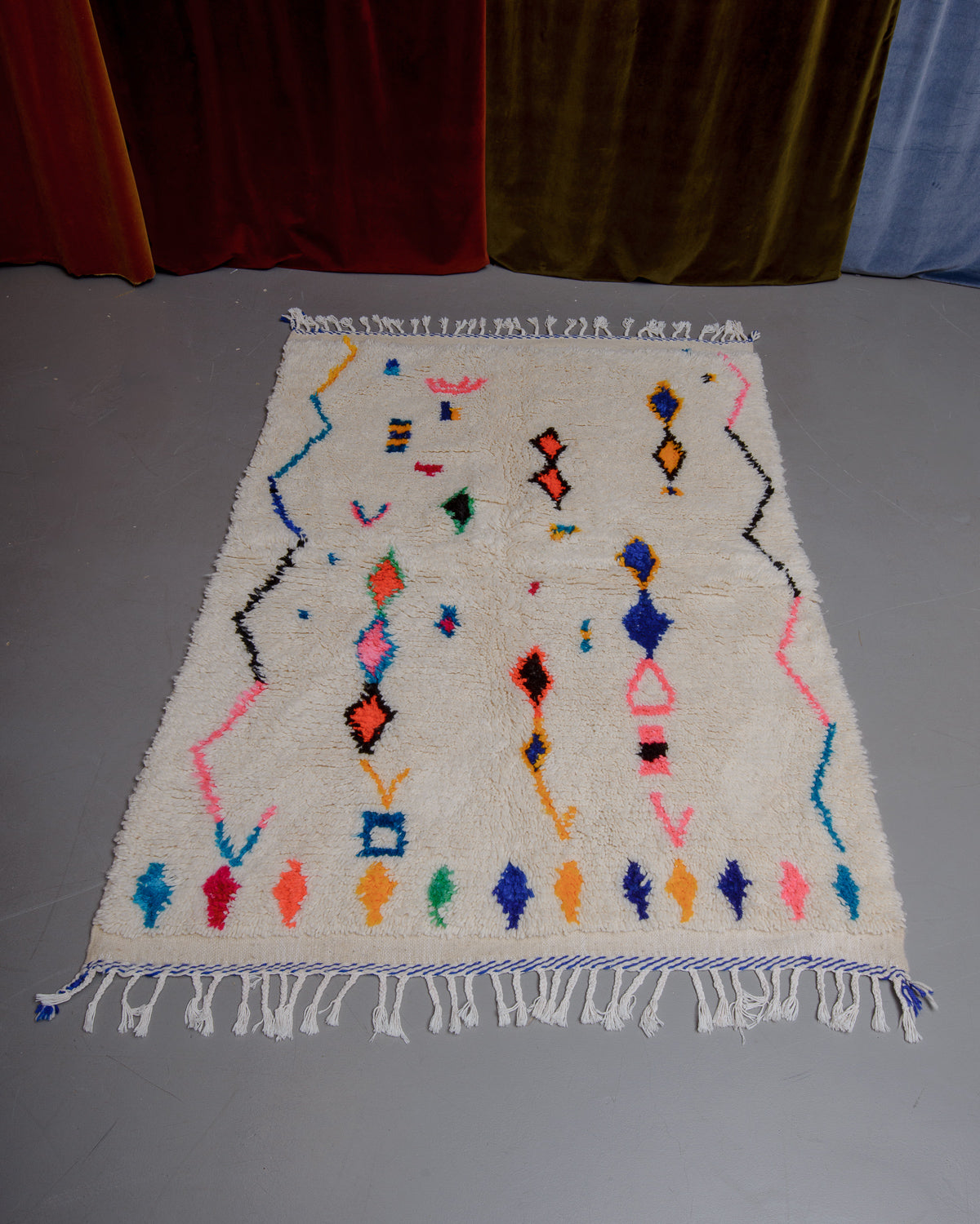 Azilal rug with colourful, abstract design laid flat on grey floor in front of a wall of velvet curtains.