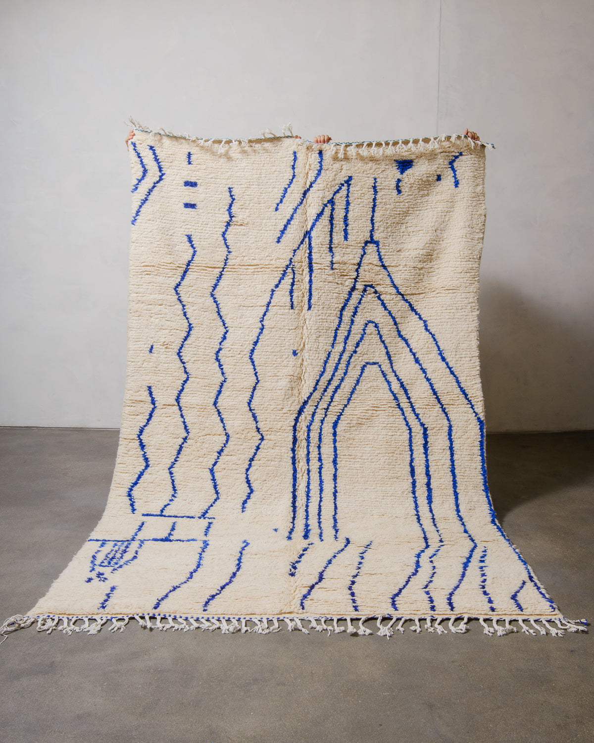 Azilal rug in cream with blue line design being held up by hands in front of a white wall. 
